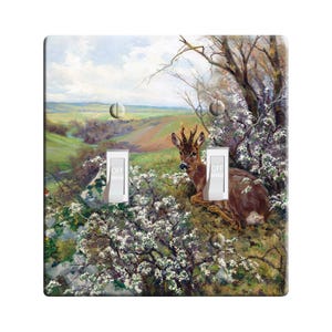May include: A light switch cover featuring a painting of a deer in a forest setting. The deer is brown with white spots and is sitting in a clearing surrounded by green trees and white flowers. The switch cover has two toggle switches labeled "OFF".