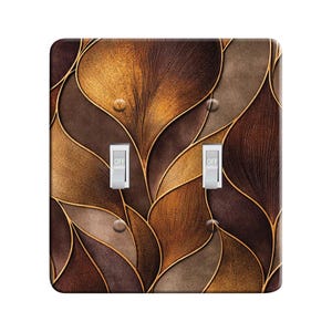May include: A decorative double light switch cover with a leaf-like pattern in shades of brown and gold. The design features curved lines and a textured appearance, with two silver switches labeled "OFF".