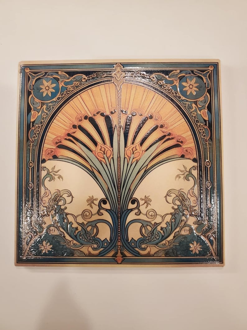 May include: Square ceramic tile with an Art Nouveau design. The tile features a central floral motif in shades of orange, blue, and green, framed by a decorative border. The design is symmetrical and detailed, with a glossy finish.