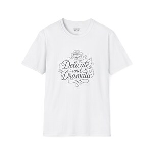 May include: White t-shirt with the text "Delicate and Dramatic" in a decorative script, accompanied by a floral design. The shirt also features the brand name "Aurora Elle" at the neckline. A casual, everyday wear item.