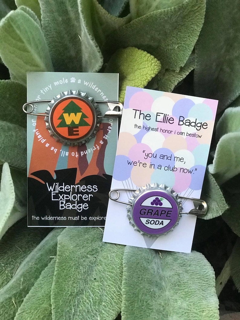 May include: Two bottle cap badges on safety pins. One features a tree design with the letters "WE" and the words "Wilderness Explorer Badge". The other says "GRAPE SODA" with a grape illustration. Both are on cards.