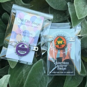 May include: Two pin-backed badges in clear plastic packaging. One features a purple "Grape Soda" design, the other a "Wilderness Explorer Badge" design with a tree. Text on the packaging reads "The Ellie Badge" and "Wilderness Explorer Badge".