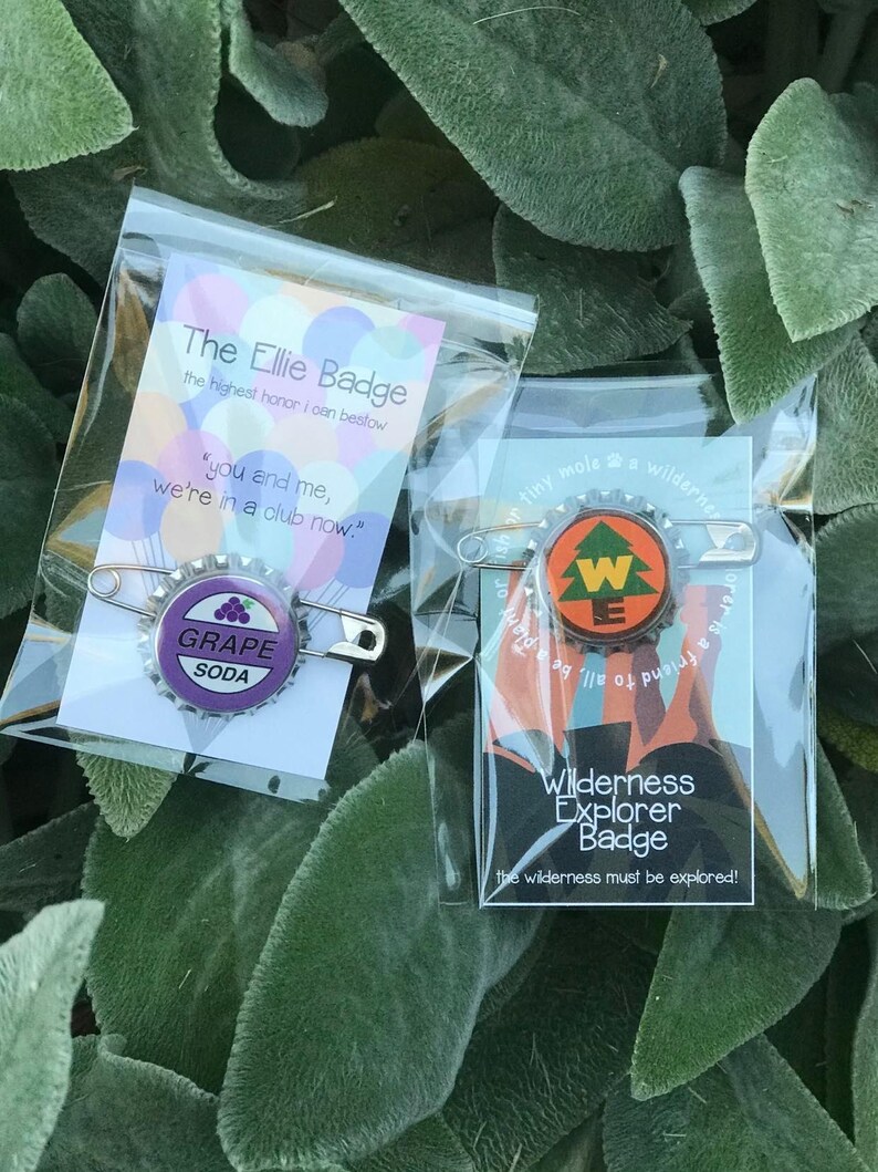May include: Two pin-backed badges in clear plastic packaging. One features a purple "Grape Soda" design, the other a "Wilderness Explorer Badge" design with a tree. Text on the packaging reads "The Ellie Badge" and "Wilderness Explorer Badge".