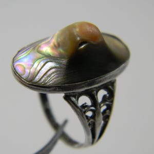 Könnte beinhalten: Silberring mit einer ovalen, irisierenden Abalone-Schale und einem erhabenen, hellbraunen Zentrum. Der Ring hat ein verziertes, durchbrochenes Design am Band.