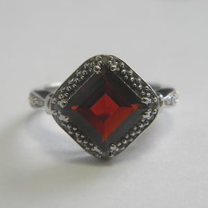 May include: A silver ring with a large, square-cut, deep red gemstone. The gemstone is set within a beaded border. The ring's band has a delicate, textured design on either side of the gemstone.