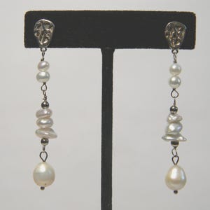 May include: A pair of dangling earrings with a silver-toned top and a cascade of white pearls. The earrings feature a mix of round and irregular-shaped pearls, culminating in a large, teardrop-shaped pearl at the bottom. The earrings are displayed on a black stand.