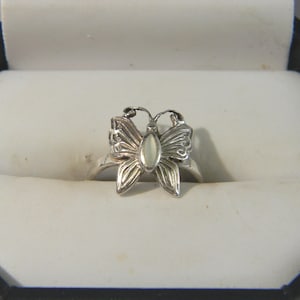 May include: A silver butterfly ring, featuring intricate wing detailing and a central oval stone. The ring is displayed in a white box with a black frame, emphasizing the jewellery's design and presentation.