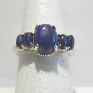 Könnte beinhalten: Ein silberner Ring mit fünf ovalen, tiefblauen Lapislazuli-Steinen. Der zentrale Stein ist größer, flankiert von zwei kleineren Steinen auf jeder Seite. Der Ring wird auf einer weißen Oberfläche präsentiert, die die polierten Edelsteine hervorhebt.