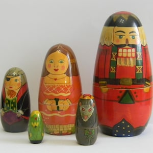 May include: A set of five wooden nesting dolls, also known as Matryoshka dolls. The largest doll is a nutcracker figure in red and gold. Other dolls depict a pirate, a woman in an orange dress, a green parrot, and a mouse. The dolls are painted with intricate details.