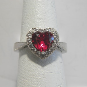 May include: A heart-shaped ring with a large, vibrant red gemstone at its center. The gemstone is surrounded by a halo of smaller, clear stones. The ring's band is silver-colored and features delicate detailing. The ring is displayed on a white surface.