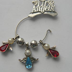 Op de afbeelding: Zilverkleurige oormanchet met de tekst "Lil Angels". De manchet heeft drie bungelende bedels: twee met rode druppelvormige edelstenen en &eacute;&eacute;n met een blauwe edelsteen, allemaal versierd met parelaccenten. Het ontwerp bevat zilveren engelen vleugels.