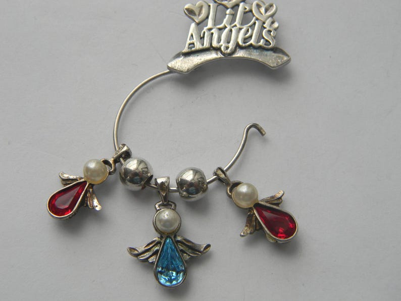 Op de afbeelding: Zilverkleurige oormanchet met de tekst "Lil Angels". De manchet heeft drie bungelende bedels: twee met rode druppelvormige edelstenen en &eacute;&eacute;n met een blauwe edelsteen, allemaal versierd met parelaccenten. Het ontwerp bevat zilveren engelen vleugels.