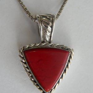 May include: A silver chain necklace with a triangular pendant featuring a red gemstone. The pendant has a silver braided border.
