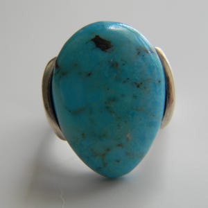 Peut inclure: Une bague en turquoise avec une bande en argent. La pierre turquoise est en forme de larme et a une couleur bleu-vert.