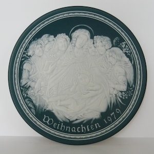 May include: A blue and white ceramic plate with a raised design of angels surrounding a baby Jesus. The plate has the text "Weihnachten 1979" around the bottom edge.