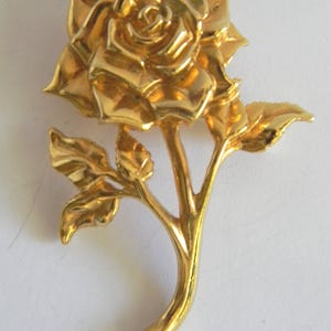May include: A gold-colored rose brooch with detailed petals and leaves. The brooch is a three-dimensional design, with the rose in full bloom and the stem curving gently. The leaves are also detailed, adding to the realistic look of the floral accessory.