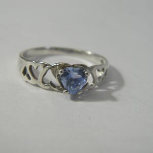 May include: A silver ring with a heart-shaped, light blue gemstone. The band has heart-shaped cutouts. The ring is set against a white background, highlighting its design and colour.