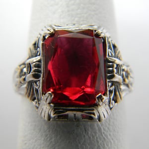 Op de afbeelding: Een zilveren ring met een sierlijk ontwerp heeft een grote, rode, rechthoekige edelsteen.