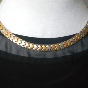 Può includere: Una collana intrecciata tricolore nei toni oro, argento e oro rosa. La collana è esposta su un busto di manichino bianco, su un top nero.