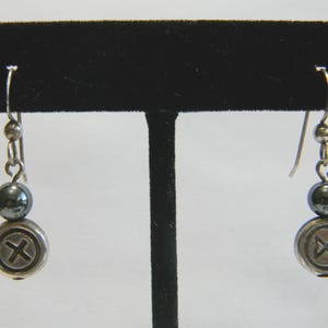 May include: Silver-tone dangle earrings. Each earring features a small silver ball, a dark gray bead, and a round charm with an X design. The earrings are displayed on a black stand against a white background. The earrings are a jewelry item.