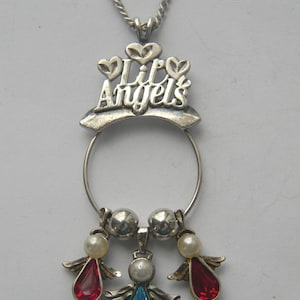 Op de afbeelding: Zilverkleurige kettinghanger met de tekst "Lil Angels". De hanger heeft een cirkelvormig ontwerp met drie engelbedels. Twee engelen hebben rode druppelvormige edelstenen en &eacute;&eacute;n heeft een blauwe edelsteen. Elke engel heeft parelaccenten.
