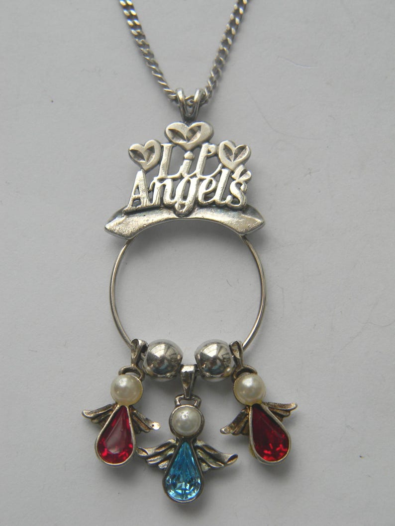 Op de afbeelding: Zilverkleurige kettinghanger met de tekst "Lil Angels". De hanger heeft een cirkelvormig ontwerp met drie engelbedels. Twee engelen hebben rode druppelvormige edelstenen en &eacute;&eacute;n heeft een blauwe edelsteen. Elke engel heeft parelaccenten.