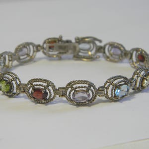 May include: Silver-toned bracelet featuring oval-shaped gemstones in various colors, including red, green, blue, and purple. The gemstones are set within a decorative, openwork design. The bracelet is secured with a clasp.