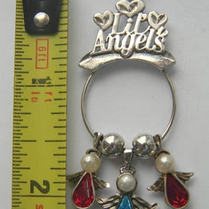 Op de afbeelding: Zilverkleurige hangerketting met de tekst "Lil Angels" en drie engelbedels. De bedels hebben rode en blauwe druppelvormige edelstenen, imitatieparels en zilveren vleugels. De ketting is ongeveer 5 cm lang.