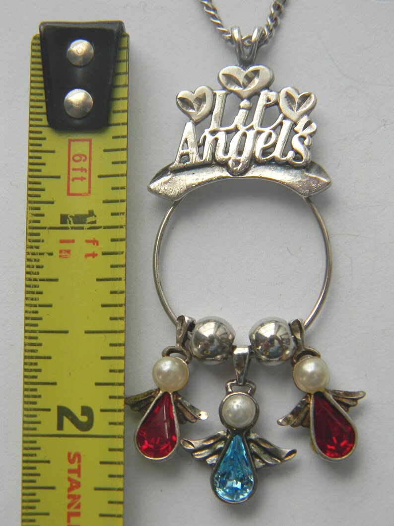 Op de afbeelding: Zilverkleurige hangerketting met de tekst "Lil Angels" en drie engelbedels. De bedels hebben rode en blauwe druppelvormige edelstenen, imitatieparels en zilveren vleugels. De ketting is ongeveer 5 cm lang.