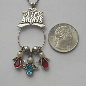 Op de afbeelding: Zilverkleurige hangerketting met de woorden "Lil Angels" en drie engelbedels. Elke engelbedel heeft een parelmoer kralenhoofd en een edelsteenlichaam in rood of blauw. Een Amerikaanse kwartdollar ligt naast de hanger.