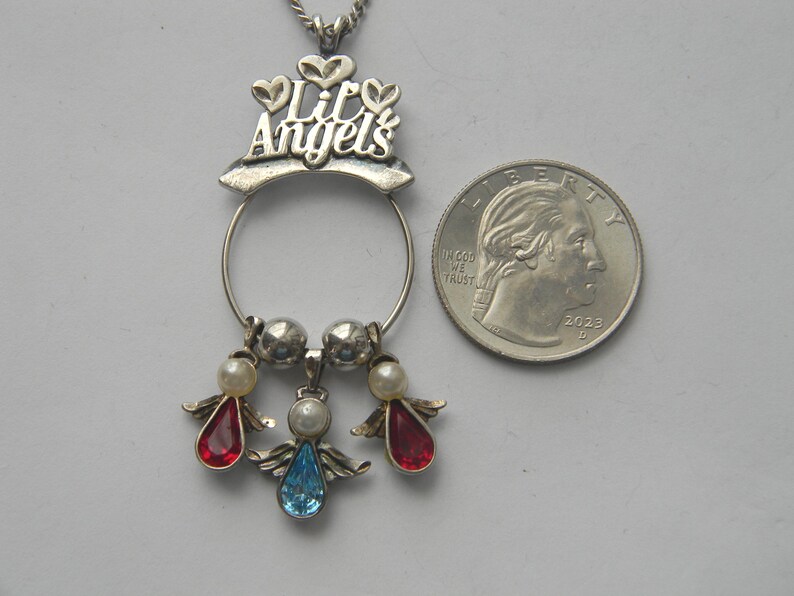 Op de afbeelding: Zilverkleurige hangerketting met de woorden "Lil Angels" en drie engelbedels. Elke engelbedel heeft een parelmoer kralenhoofd en een edelsteenlichaam in rood of blauw. Een Amerikaanse kwartdollar ligt naast de hanger.