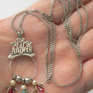 Op de afbeelding: Zilverkleurige ketting met een hanger met de tekst "Lily Angels" en drie engelbedels. De bedels hebben rode, blauwe en parelkleurige accenten. De ketting heeft een fijne ketting.