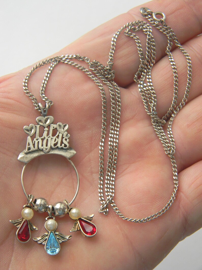 Op de afbeelding: Zilverkleurige ketting met een hanger met de tekst "Lily Angels" en drie engelbedels. De bedels hebben rode, blauwe en parelkleurige accenten. De ketting heeft een fijne ketting.
