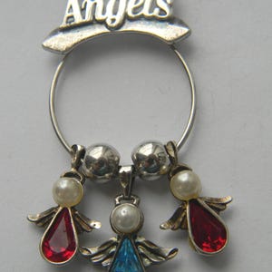 Op de afbeelding: Een zilverkleurige bedel met de tekst "Lit Angels" boven een ring. De bedel heeft drie engelhangers met parelaccenten en rode en blauwe druppelvormige edelstenen. De engelen hebben zilveren vleugels.