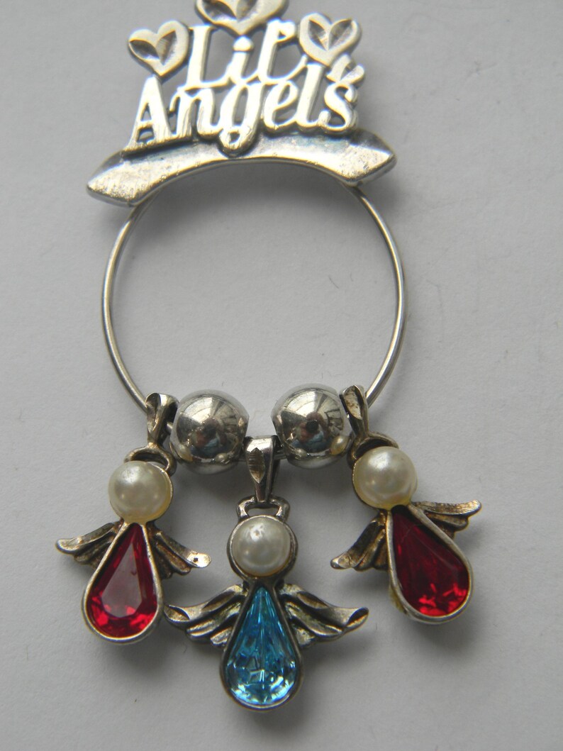 Op de afbeelding: Een zilverkleurige bedel met de tekst "Lit Angels" boven een ring. De bedel heeft drie engelhangers met parelaccenten en rode en blauwe druppelvormige edelstenen. De engelen hebben zilveren vleugels.