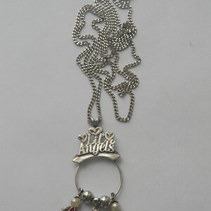 Op de afbeelding: Zilverkleurige ketting met een ketting en een hanger. De hanger heeft de tekst "Lil' Angels" boven een cirkelvormig frame. Onder het frame bevinden zich drie engelbedels, elk met een gekleurde edelsteen: rood, blauw en helder.