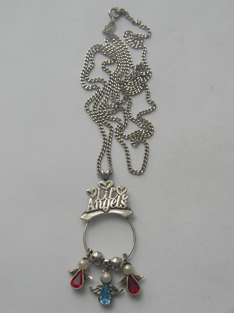 Op de afbeelding: Zilverkleurige ketting met een ketting en een hanger. De hanger heeft de tekst "Lil' Angels" boven een cirkelvormig frame. Onder het frame bevinden zich drie engelbedels, elk met een gekleurde edelsteen: rood, blauw en helder.