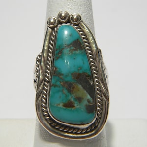 Op de afbeelding: Een zilveren ring met een grote, ovale turkoois steen. De ring heeft een gedetailleerd ontwerp met touwachtige randen en decoratieve elementen. De turkoois steen heeft een levendige blauwgroene kleur.