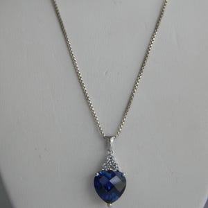 Pode incluir: Um colar de corrente de prata com um pingente em forma de coração. O pingente apresenta uma grande gema azul cercada por gemas transparentes menores.