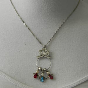 Op de afbeelding: Zilverkleurige ketting met een hanger met de tekst "My Angels". De hanger heeft een cirkelvormig ontwerp met drie kleine engelenfiguren, elk met een rode, blauwe en parelkleurige edelsteen. De ketting wordt gepresenteerd op een witte achtergrond.