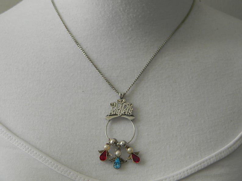 Op de afbeelding: Zilverkleurige ketting met een hanger met de tekst "My Angels". De hanger heeft een cirkelvormig ontwerp met drie kleine engelenfiguren, elk met een rode, blauwe en parelkleurige edelsteen. De ketting wordt gepresenteerd op een witte achtergrond.