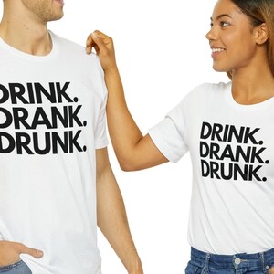 Op de afbeelding: Twee witte T-shirts met zwarte tekst die "DRINK. DRANK. DRUNK." zegt.