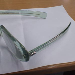 May include: A pair of translucent light green eyeglasses with a rectangular frame shape. The glasses feature thin, matching green temples and clear lenses, positioned at an angle to show the side profile and the front frame structure clearly.