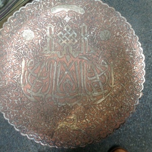 May include: A round, copper tray with intricate silver filigree designs and Arabic calligraphy. The tray has a scalloped edge and a worn, antique finish.