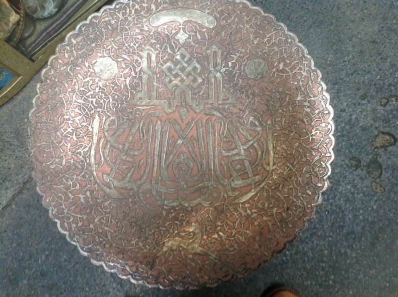 May include: A round, copper tray with intricate silver filigree designs and Arabic calligraphy. The tray has a scalloped edge and a worn, antique finish.