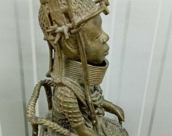 Op de afbeelding: Een gouden metalen sculptuur van een zittend figuur met een gedetailleerd hoofdtooi en ingewikkelde patronen over de hele oppervlakte. Het figuur zit op een geweven stoel met een vergelijkbaar ingewikkeld ontwerp.