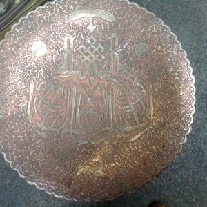 May include: A round copper tray with intricate silver filigree designs and Arabic calligraphy. The tray has a scalloped edge and a worn, antique look.