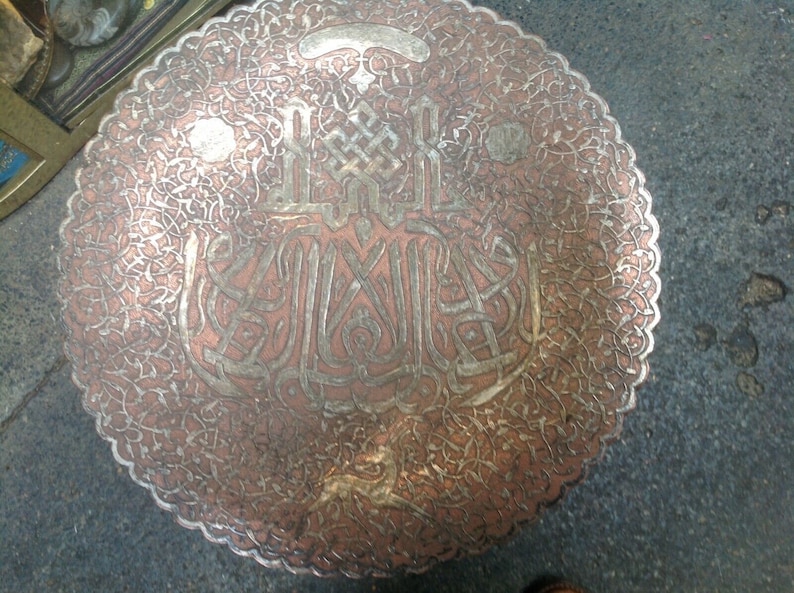 May include: A round copper tray with intricate silver filigree designs and Arabic calligraphy. The tray has a scalloped edge and a worn, antique look.