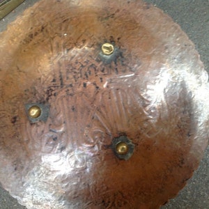 May include: A copper plate with a decorative design and four brass accents. The plate is round and has a scalloped edge.