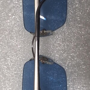 May include: Rectangular eyeglasses featuring translucent blue lenses and a thin, metallic silver-toned frame. The design includes a straight bridge, adjustable clear nose pads, and slender temples that extend from the outer corners of the frames.